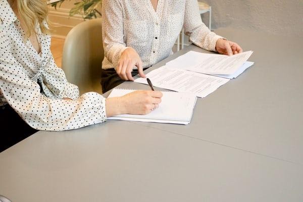 Two women sat at a table, one with a pen in hand about to sign a document. 