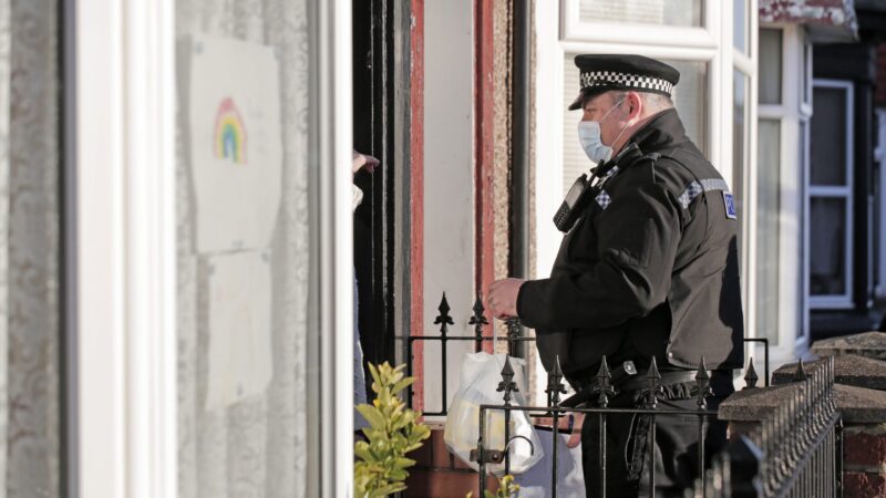 A police officer in uniform stands on a doorstep speaking to an unseen resident